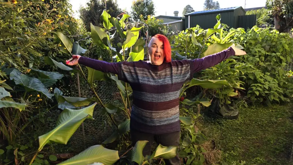 Woman stands in a subtropical garden, surrounded by lush foliage, arms outstretched and looking happy and excited