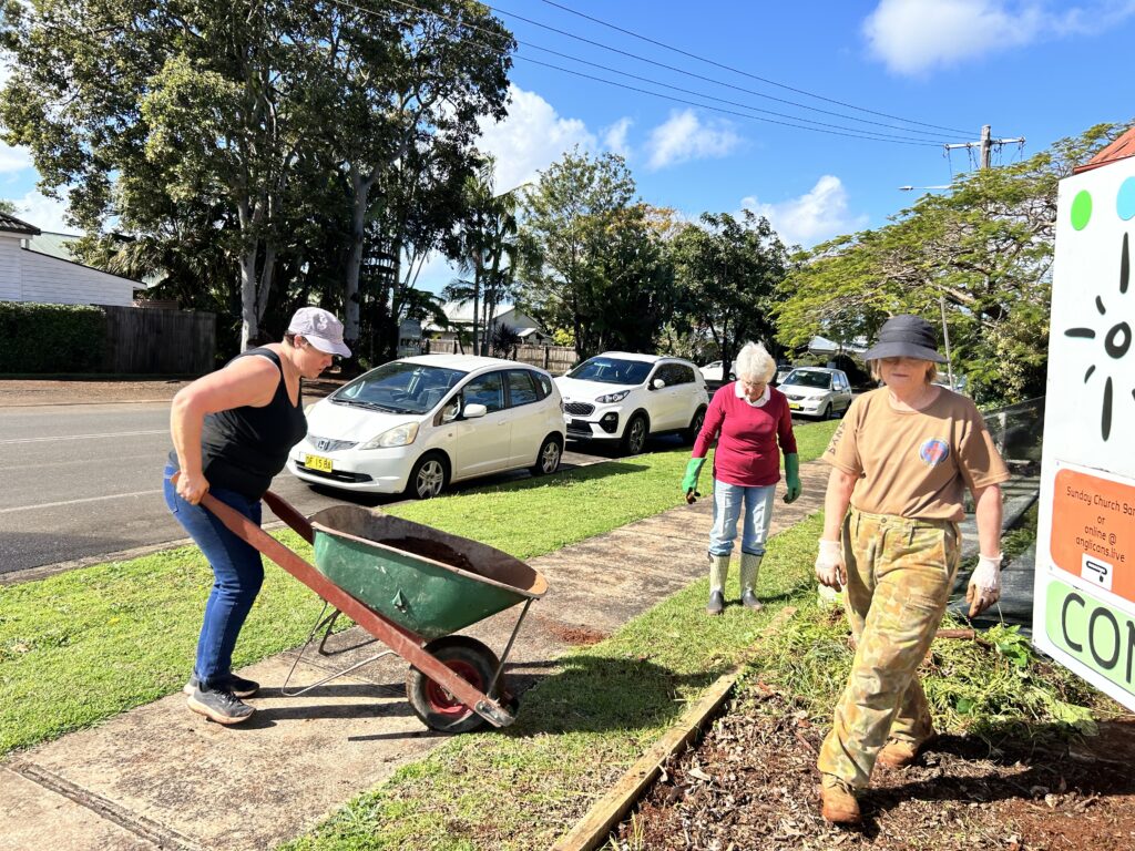 Woman pushes wheelbarrow across footpath while other gardeners walk through verge garden 